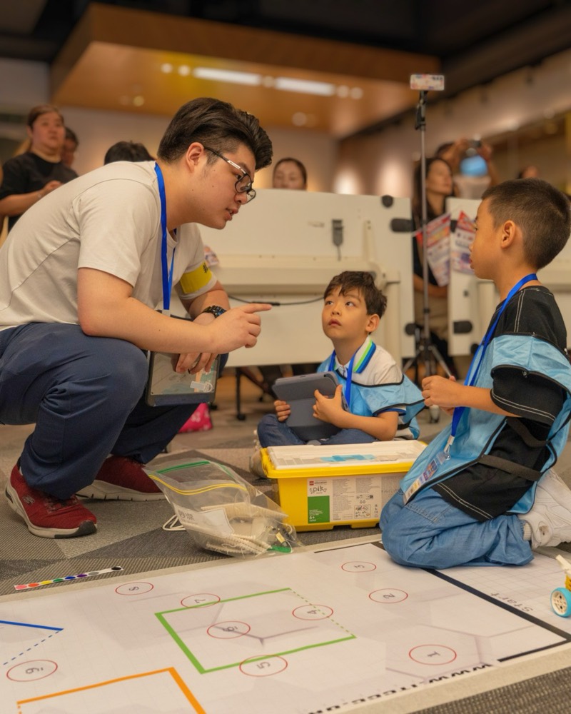 A JBC mentor kneels to guide young competitors at the game table — guidance over judgment