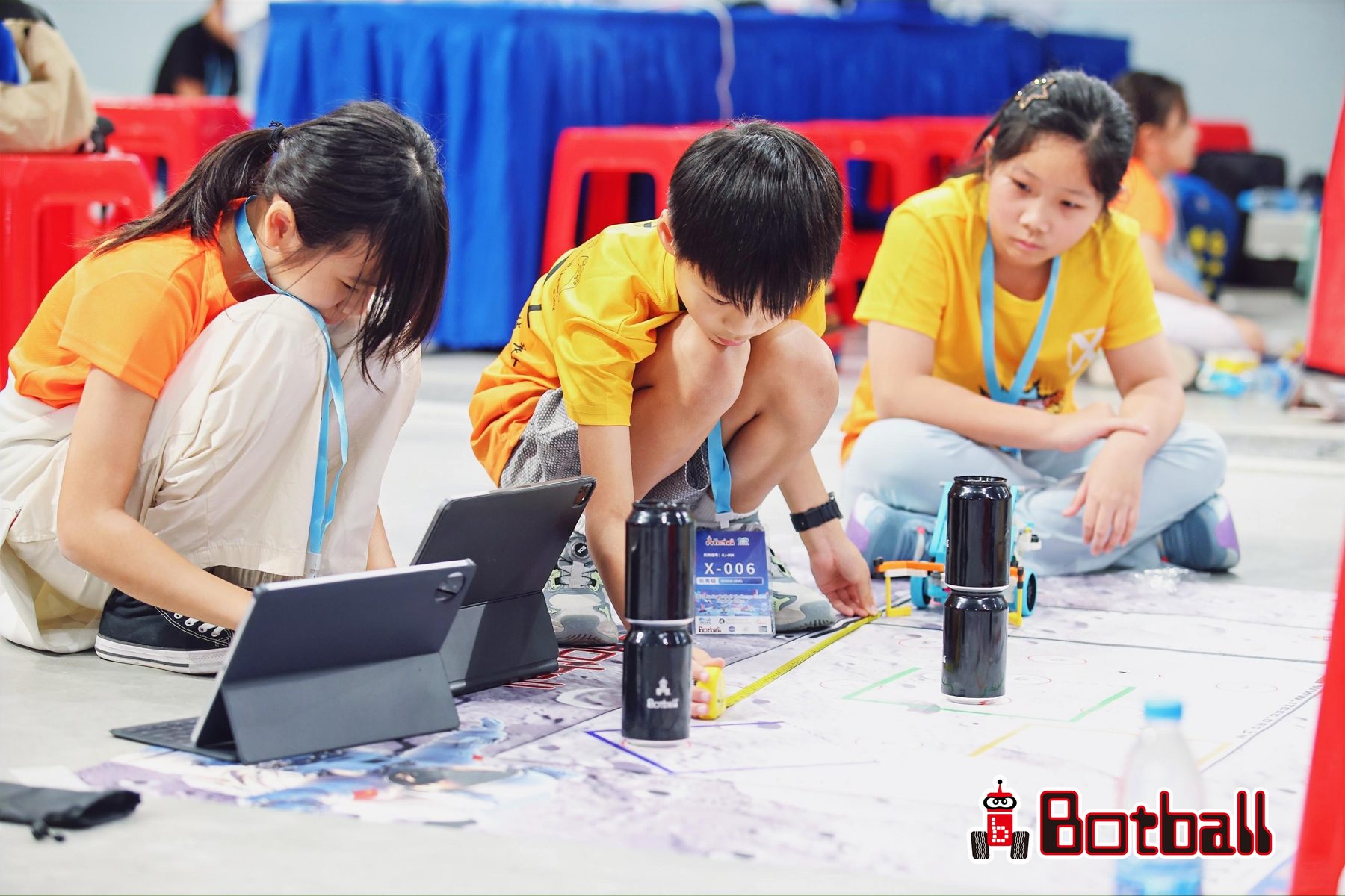 Students measure their robot's run on the JBC game mat while a teammate checks the program on a tablet